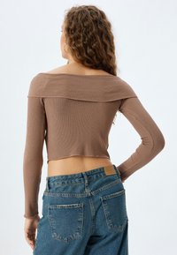 Brun, ribbet off-the-shoulder cropped top med lange ærmer, parret med højtaljede blå denim jeans. Har en rå kant og tætsiddende design.