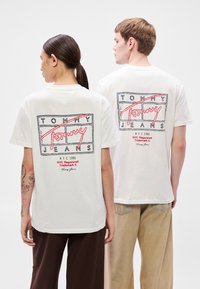 Valkoiset puuvillaiset t-paidat, joissa on punainen ja musta graafinen logo takana, pyöreä pääntie ja lyhyet hihat.