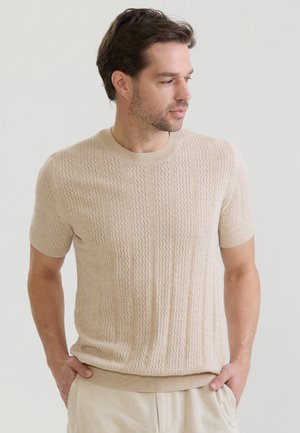 Homme aux cheveux châtain clair portant un pull en maille beige à manches courtes et un pantalon clair, regardant légèrement vers sa droite avec les mains dans les poches.
