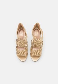 Sandales à talons hauts dorées avec des brides croisées et un détail tissé au niveau des orteils, dotées d'une finition lisse et brillante et d'une fermeture éclair à l'arrière.