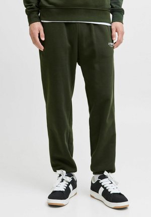 Grønne sweatpants lavet af blødt stof, med elastisk talje og et lille hvidt logo. Bærer sorte og hvide sneakers.