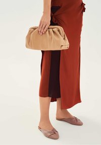 Kobieta ubrana w spódnicę w kolorze rdzy i fioletowe wsuwane baleriny, trzyma beżową kopertówkę z marszczeniami przy boku.