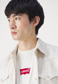 Bežinė džinsinio audinio marškinėliai virš baltos marškinėlių su raudonu Levi's logotipu. Artimas vaizdas į viršutinę kūno dalį, akcentuojantis apykaklės detales ir audinio tekstūrą.