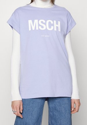 Personne portant une chemise lavande à manches courtes avec le texte « MSCH EST MMX » sur un col roulé blanc côtelé et un jean bleu.