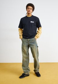 Musta lyhythihainen t-paita kerroksittain beige pitkähihaisen paidan päällä, yhdistettynä vaaleansinsisiin löysiin farkkuihin ja mustiin tennareihin. Yksinkertainen logotyyli.