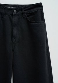 Jeans de mezclilla negra con cinturilla estándar, bolsillos frontales y un solo botón en la parte delantera. Diseño minimalista con una textura suave.