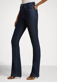 Tmavomodré rifle z denimu s rozšírenými nohavicami, vysokým pásom, viditeľným prešitím a prednými vreckami. Dopĺňajú ich hnedé sandále so remienkami.