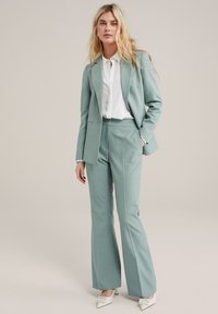Traje ajustado de color verde claro con americana de un solo botón y pantalones acampanados, combinado con una camisa blanca de botones y tacones color crema.