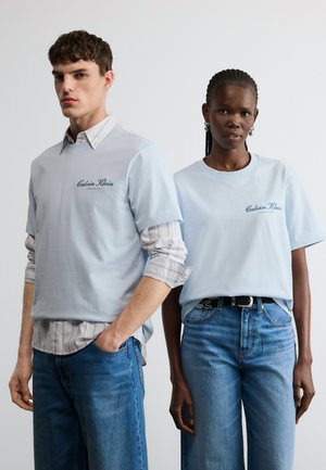 Két modell világoskék Calvin Klein pólót és kék farmert visel, egyszerű fehér háttér előtt állnak.