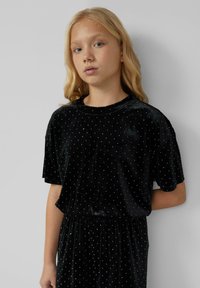 Zwarte fluwelen jurk met korte mouwen en witte stippen, met een elastische tailleband. Zachte textuur en casual silhouet.