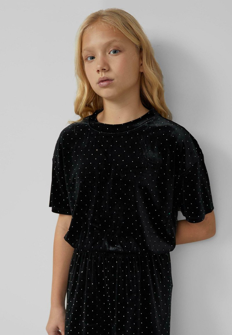 Zwarte fluwelen jurk met korte mouwen en witte stippen, met een elastische tailleband. Zachte textuur en casual silhouet.