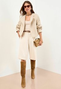 Vrouw in een beige cropped jack, mouwloze top, knielange shorts, bruine suede kniehoge laarzen, zonnebril en met een gestructureerde handtas.