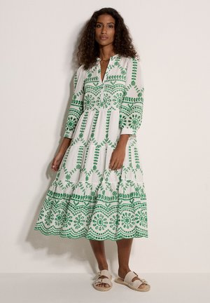 Mujer vestida con un vestido midi blanco con bordados florales verdes y mangas abullonadas, combinando con sandalias beige de doble tira, parada contra una pared blanca.