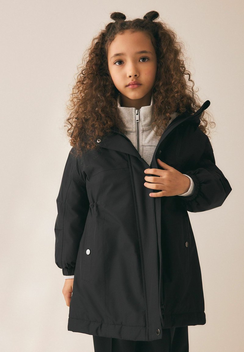 Joven niña con pelo rizado que lleva un abrigo de invierno negro sobre un suéter gris claro con cremallera, con un fondo liso.