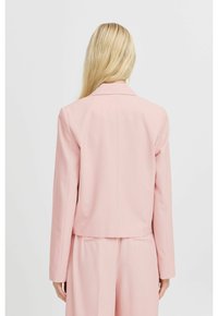 Světle růžové sako na míru s vykrojeným límcem, strukturovanými rameny a zkráceným střihem. Hladká textura a minimalistické detaily.