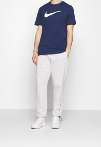 Henkilö, jolla on yllään tummansininen Nike-t-paita, vaaleanharmaat verkkohousut, joissa teksti vasemmassa lahkeessa, ja valkoiset lenkkitossut, seisoo valkoista taustaa vasten.