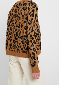Brązowy sweter z czarno-szarym wzorem w panterkę, z prążkowanymi mankietami i dołem, wykonany z dzianiny, noszony na kremowych spodniach z kordurowej tkaniny.