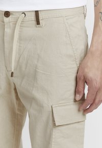 Pantalons cargo beige en tissu texturé, avec taille à cordon de serrage, poche latérale et détail de bouton.