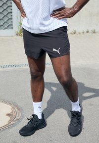 Czarne sportowe szorty z logo Puma, w połączeniu z czarnymi butami sportowymi i białymi skarpetami. W tle asfalt i minimalna zieleń.