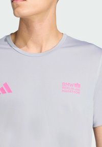 Kratak prikaz osobe koja nosi svijetlosivu Adidas majicu s jarko ružičastim logotipom BMW Berlinskog maratona i Adidas potezima na prsima.