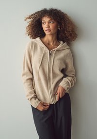 Felpa beige con zip e cappuccio regolabile, polsini e orlo a costine. Tessuto morbido con una leggera texture, abbinata a pantaloni neri.