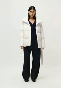 Kvinne med rett, mørkt hår har på seg en hvit boblejakke over en marineblå skjorte med knapper, svarte vide bukser og svarte sko, står med en nøytral holdning.