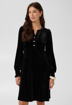 Femme aux longs cheveux bruns portant une robe noire en velours à manches longues avec des boutons en perles et un bracelet beige, debout devant un fond uni.