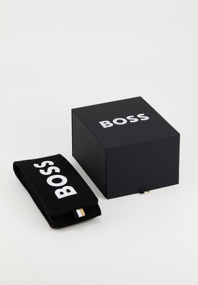 Boîte noire à finition mate avec le logo blanc "BOSS", à côté d'une pochette noire avec le même logo et un petit accent doré.