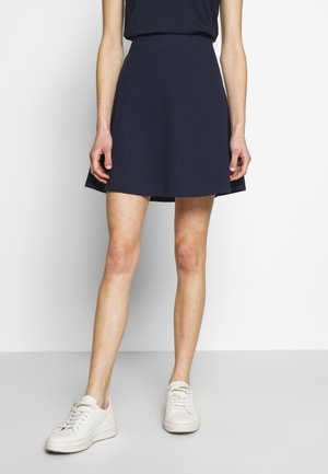 Vrouw met een navy blauwe A-lijn rok, zwart shirt en witte sneakers, staand tegen een effen witte achtergrond.