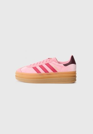 Roze suède sneakers met drie donkeroze strepen, een ronde neus en een dikke gumzool. Het woord "GAZELLE" is in het goud op de zijkant gegraveerd.