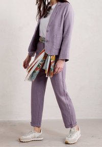 Veste blazer en lin violet à deux boutons et poches avant, associée à un pantalon assorti. Accessoirisée d'une ceinture à motif floral et de baskets claires.