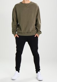 Sudadera verde oliva de manga larga y dobladillo acanalado, combinada con joggers negros de corte ajustado, usados con zapatillas blancas.