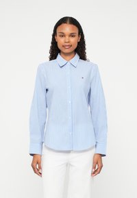 Light blue striped shirt with a button-down collar, long sleeves, and a small logo accent on the chest. Soft fabric and tailored shape.