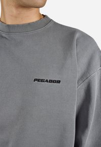 Grauer Sweatshirt mit geripptem Kragen und Bündchen, auf dem die Worte "PEGADOR" in schwarzem Stickerei auf der Brust stehen. Weicher, strukturierter Stoff.