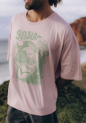 Jasnoróżowa koszulka z zielonym nadrukiem przedstawiającym syrenę otoczoną morskimi elementami i tekstem. Krótkie rękawy i luźny krój.