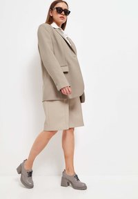 Blazer beige ajustado sobre una camisa blanca, combinado con una falda beige. Zapatos de tacón grises con cordones negros y suelas texturizadas.