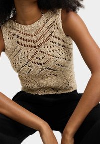 Beige gehaakt mouwloos topje met ingewikkelde openwerkpatronen, textuurgebreide stof en een hoge halslijn. In combinatie met zwarte broek.