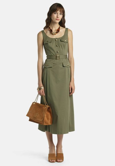 Sieviete tērpusies olīvu zaļā, bezroku, vidēja gara kleitā ar jostu un priekšpusē kabatām, rokās tur brūnu zamšādas rokassomiņu un valkā saskaņotas mūļu kurpes un biezas kaklarotas.