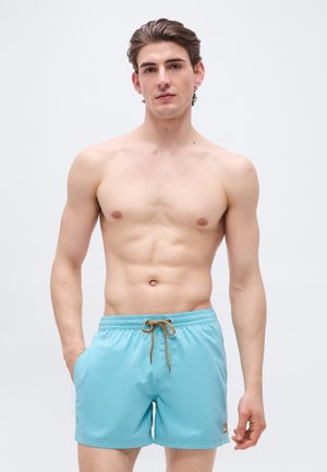 Jonge man staand, zonder shirt, draagt lichtblauwe zwembroek met bruine trekkoord, tegen een effen lichte achtergrond.