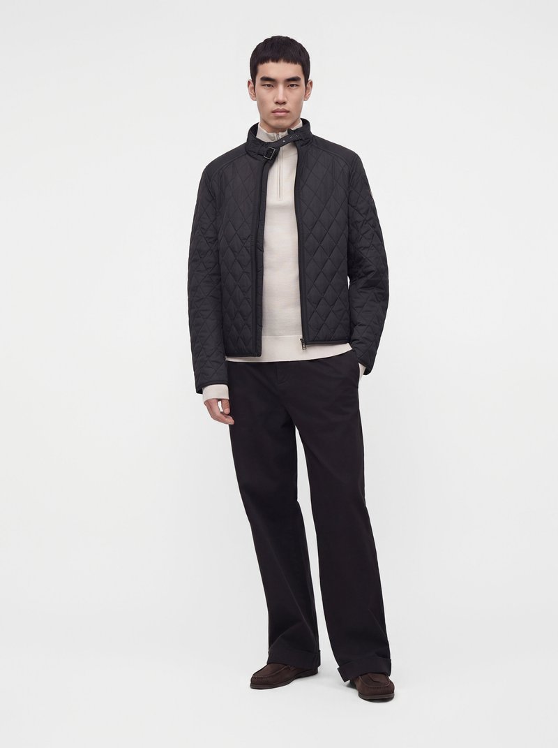 Modèle masculin portant une veste matelassée noire sur un pull beige, un pantalon noir et des chaussures marron, debout devant un fond clair uni.