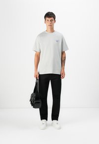 Helles graues Baumwoll-T-Shirt mit kurzen Ärmeln und einem kleinen blauen Grafikdesign. Kombiniert mit schwarzen Hosen und weißen Turnschuhen, hält eine schwarze Tasche.