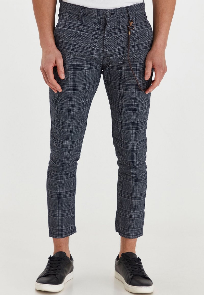Pantalon slim gris à carreaux coupé à la cheville avec un accessoire chaîne marron, porté avec des baskets noires sur une personne debout.