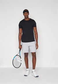 Μαύρο κοντομάνικο μπλουζάκι, γκρι σορτς, κρατώντας μία ρακέτα τένις. Φοράει λευκά sneakers με μπλε λεπτομέρειες και μία μαύρη κορδέλα στο κεφάλι.