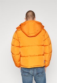 Giacca a vento arancione con cappuccio, caratterizzata da un design trapuntato, una texture lucida e polsini elastici. Abbinata a jeans di denim blu.