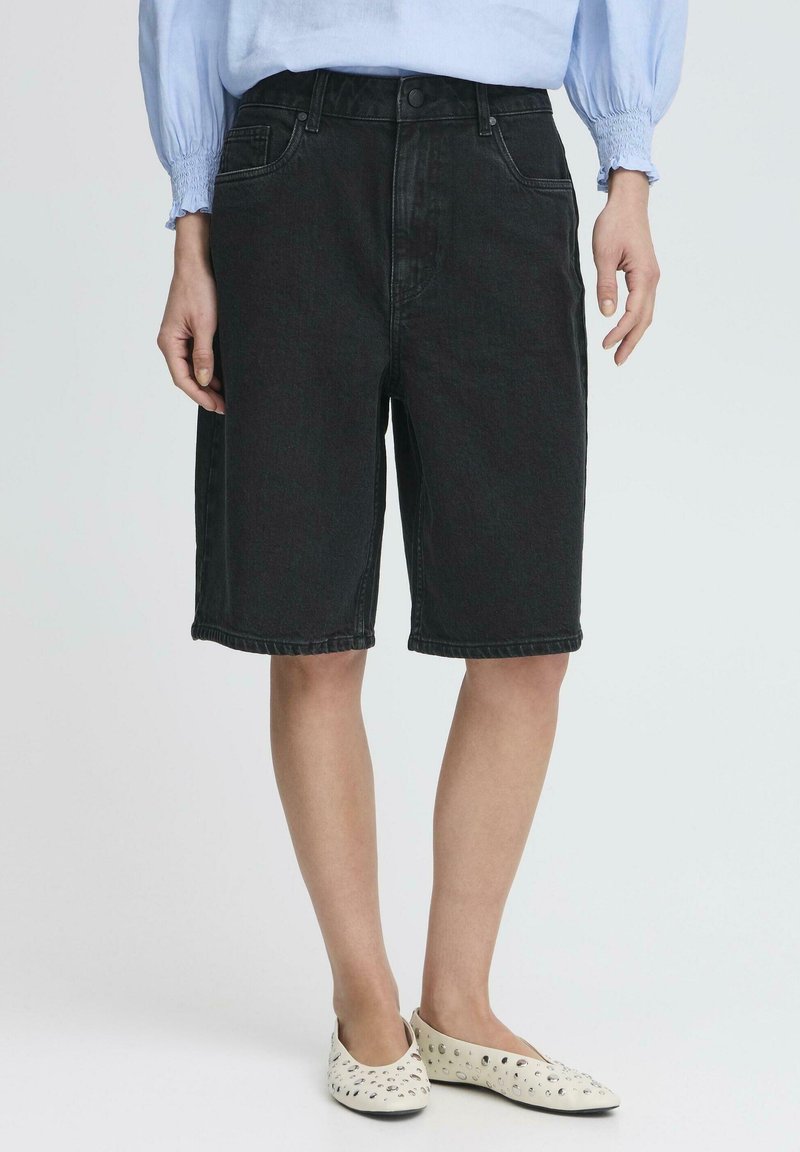Femme portant un short en jean noir à hauteur des genoux, un chemisier bleu clair à manches longues et des ballerines blanches perforées, debout sur fond blanc.