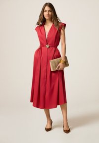 Rotes ärmelloses Kleid mit V-Ausschnitt, tailliert mit Gürtel und plissiertem Rock. Das Model hält eine beigefarbene geflochtene Clutch und trägt Leopardenmuster-High Heels.