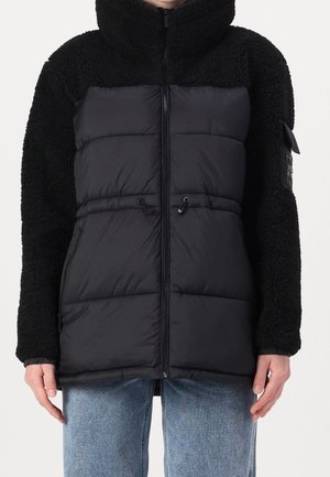 Osoba ubrana w czarną kurtkę z kapturem, z rękawami z polaru i pikowanym przodem, w połączeniu z jasnoniebieskimi dżinsami, stojąca na jednolitym tle.