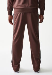 Καφέ παντελόνια φόρμας από λείο ύφασμα, με πλευρικές ρίγες σε μαύρο και άσπρο. Ελαστική ζώνη μέσης και μανσέτες, με λευκές λεπτομέρειες στα παπούτσια.