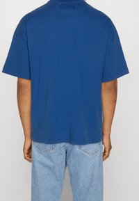 Blaues Baumwoll-T-Shirt mit lockerer Passform, kurzen Ärmeln und rundem Halsausschnitt. Die Rückseite ist schlicht mit minimalen Nähdetails.