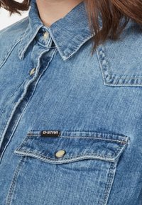 Långärmad denimskjorta i ljusblått med spetsig krage, knappstängning, bröstficka med en svart etikett och subtil textur.